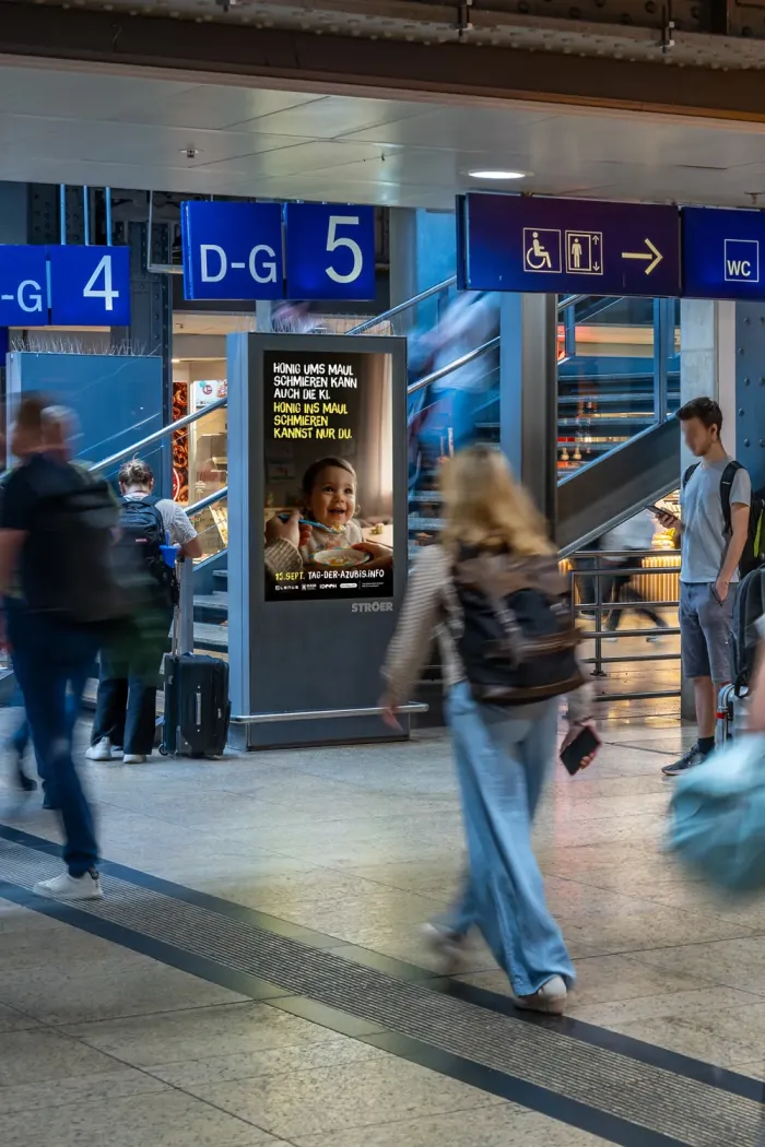 Digitales City-Light-Poster in einer Bahnhofshalle zeigt das Kampagnenmotiv „Honig ums Maul schmieren kann auch die KI. Honig ins Maul schmieren kannst nur du.“
